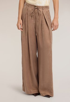 Donna con pantaloni marroni a vita alta e gamba larga con cintura annodata, in piedi davanti a uno sfondo chiaro e uniforme.