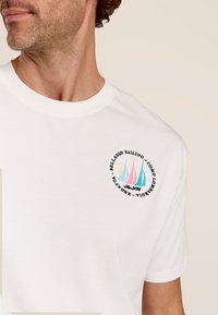 Witte katoenen t-shirt met een kleurrijke zeilgrafiek op de linkerkant van de borst, omrand met een ronde rand met tekstdetails. Korte mouwen.