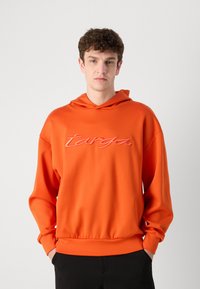 Felpa arancione con una consistenza morbida, dotata di cappuccio con coulisse e logo "targa" ricamato in rosa. Vestibilità ampia con polsini e orlo a coste.
