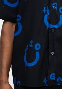 Μαύρο πουκάμισο με κοντά μανίκια, διακοσμημένο με μπλε αφηρημένα πρόσωπα-χαμόγελα και σύμβολα, μερικώς κουμπωμένο, φορεμένο από άτομο με ορατό αριστερό χέρι.