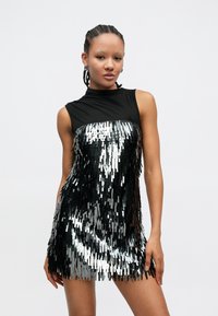 Μαύρο αμάνικο φόρεμα με εφαρμοστό πάνω μέρος και στρωματοποιημένο, λαμπερό ασημί σχέδιο με φούντες. Λείο ύφασμα με αντανακλαστικά στοιχεία σε όλο το μήκος.