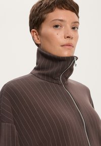 Rjava zip-up majica z visokim ovratnikom, ki ima vertikalne svetle proge, mehak material in srebrno zadrgo.