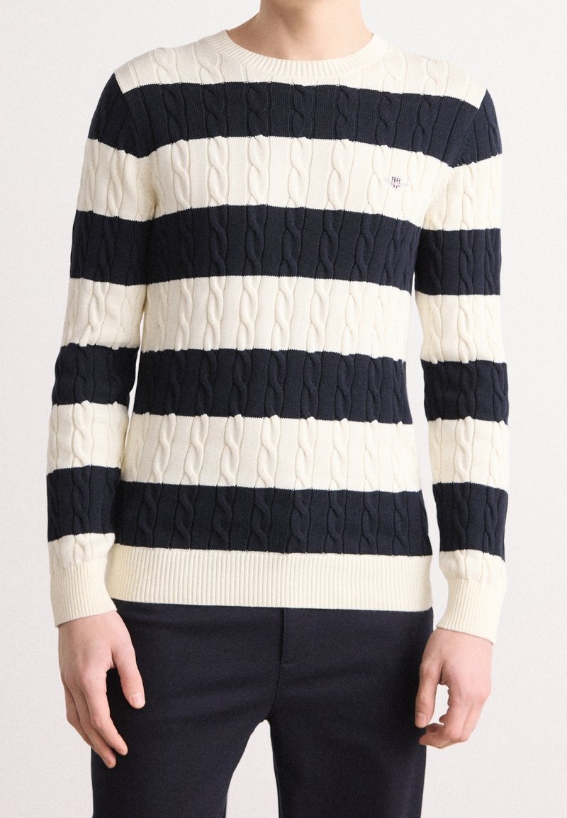 Homme portant un pull à rayures horizontales bleu marine et blanches en tricot torsadé et un pantalon foncé, les bras détendus le long du corps.