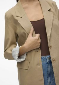 Beige kavaj med notch-krage, två knappar och en ficka. Vit och grårandig manschett syns från ärmen. Brun topp och denimjeans synliga.