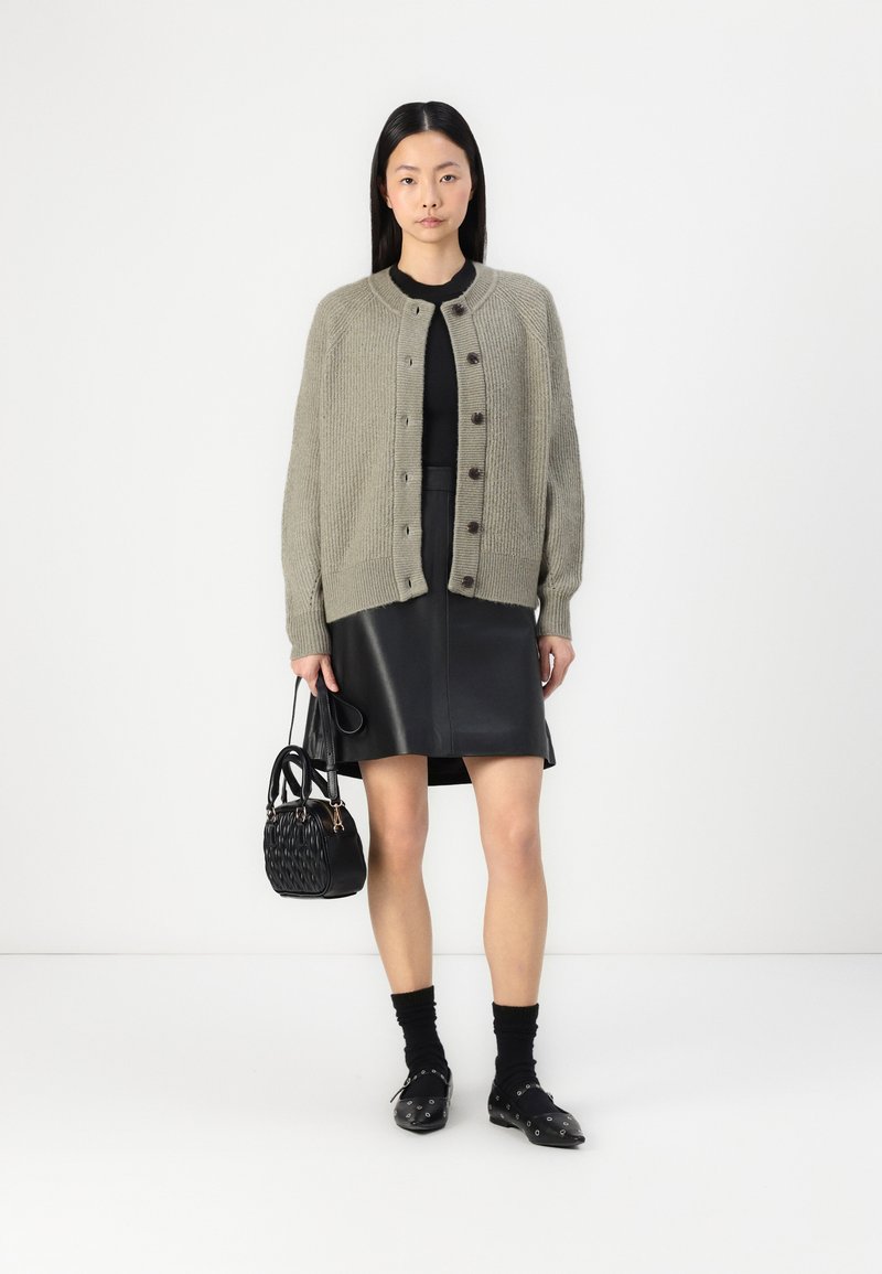 Helle olivfarbene Strickjacke mit rundem Ausschnitt und dunklen Knöpfen, kombiniert mit einem schmalen, schwarzen Lederrock und schwarzen Accessoires.