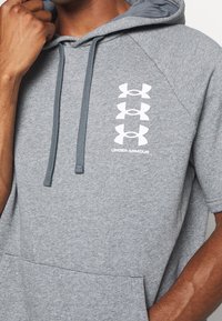 Hombre con una sudadera corta de manga gris con tres logotipos blancos de Under Armour apilados verticalmente en el pecho y cordones colgando.