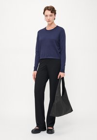 Marineblå langermet skjorte, svarte bukser og en svart skulderveske. Vesken er myk med en slouchy form. Modellen poserer mot en hvit bakgrunn.