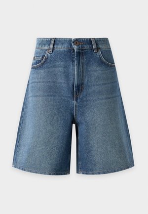 Femme portant une chemise ample à rayures boutonnée, un short en jean à hauteur du genou, et des bottes marron mi-mollet, debout avec une main dans la poche.