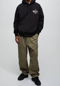 Osoba ubrana w czarną bluzę z kapturem z kieszenią i grafiką, oliwkowozielone spodnie bojówki oraz czarne trampki, stojąca na tle jednolitego jasnego tła.