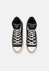 Une paire de baskets montantes en toile noires avec des lacets blancs, des embouts en caoutchouc, des œillets dorés et des crosses brodées bleu, blanc et rouge sur les languettes.