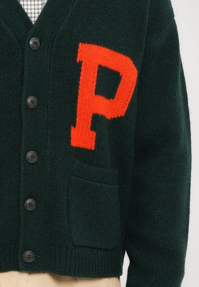 Cárdigan de punto verde oscuro con una gran letra "P" naranja, cinco botones negros, dobladillo acanalado y un bolsillo frontal.