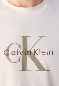 Hvidt stof med store, lyse beige "CK" bogstaver og "Calvin Klein" broderet med mindre brunt garn på tværs af midten.