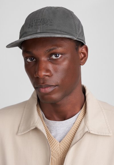 Γκρίζο καπέλο από ύφασμα, με καμπυλωτό γείσο και ανάγλυφα γραφικά στο μπροστινό μέρος, φορεμένο από ένα άτομο σε ανοιχτόχρωμο μπουφάν και πουλόβερ.