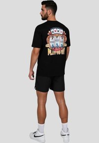 Camiseta de algodón negra con un diseño gráfico que presenta una máquina tragamonedas y cartas, con el texto "JUGAR PARA GANAR" en amarillo. Combinada con pantalones cortos negros.