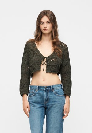 Γυναίκα που φοράει μια σκούρα πράσινη πλεκτή κοντή ζακέτα δεμένη μπροστά και μπλε ψηλόμεσα τζιν, στέκεται μπροστά από απλό φόντο.