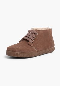 Stivali ankle in suede marrone con punta arrotondata, dotati di lacci beige, occhielli dorati e una suola in gomma marrone testurizzata.