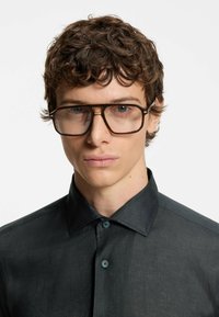 Brown oversized glasses with a gold accent, worn by a model in a textured dark shirt featuring a button-down collar and blue buttons.