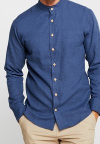 Mörkblå skjorta med ståkrage, som har träknappar och långa ärmar. Tyget verkar slätt och har en texturerad yta.