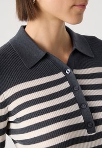 Haut en tricot à rayures gris foncé et crème avec un col à boutons, comportant quatre boutons foncés et un tissu texturé.