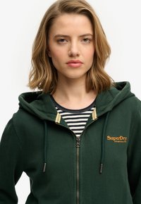Groen zip-up hoodie met capuchon, met een gele accent bij de halslijn en het logo "SuperDry Athletics" in oranje op de borst. Gestreept shirt eronder.