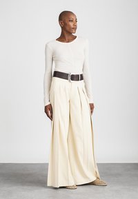 Witte langmogige top, hoog-waisted crèmekleurige plooibroek, wijd gesneden, bruine riem met een grote gesp, en beigekleurige gevlochten muiltjes.