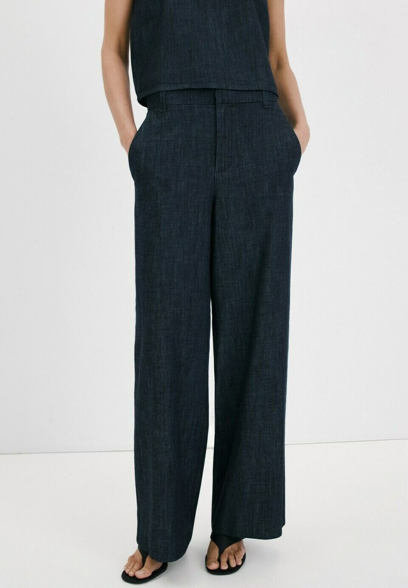 Femme portant un pantalon large en lin sombre avec des poches latérales et des sandales noires à bout ouvert, debout devant un arrière-plan clair uni.