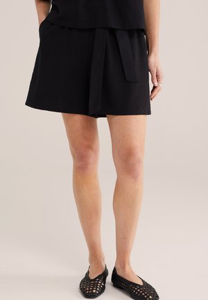 Kobieta ubrana w czarne szorty z wysokim stanem i paskiem do wiązania, czarne plecione buty wsuwane oraz czarny top na jednolitym tle.