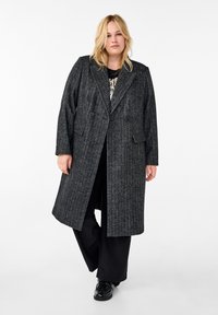 Cappotto lungo grigio in misto lana con design doppiopetto, ampi revers, tessuto testurizzato e tasche frontali. Indossato sopra un top nero e pantaloni.