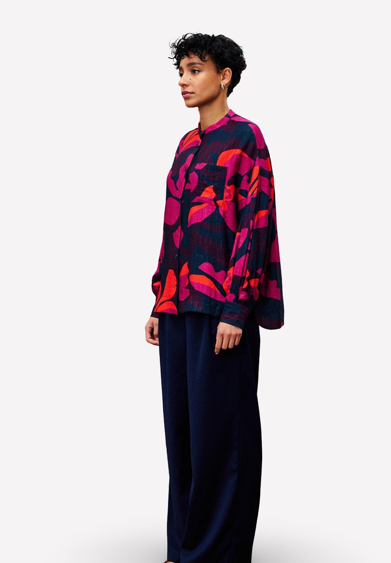 Une personne avec des cheveux bouclés courts porte une chemise à motifs floraux vibrants rouges et violets ainsi qu'un pantalon bleu foncé ample, se tenant devant un fond blanc.