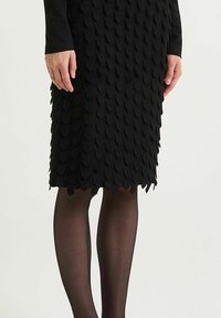 Jupe crayon noire en maille avec une texture festonnée, atteignant le milieu du mollet, associée à des collants noirs transparents. Aucun accessoire ni accent visible.