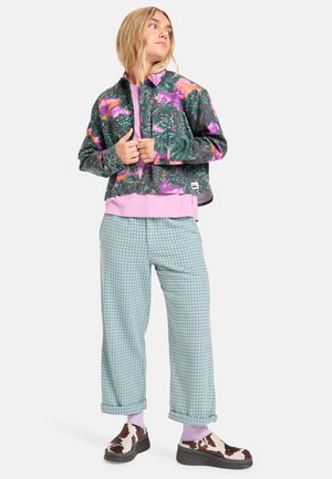 Kleurvolle korte jas met tropenprint, gelaagd over een roze shirt. Losse, lichtblauwe geblokte broek en gestreepte schoenen maken de outfit compleet.