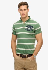 Zielona w paski koszulka polo z krótkimi rękawami, kołnierzykiem na dwa guziki, granatowym haftem i kontrastującymi kremowymi paskami. Nosi się ją z beżowymi spodniami i brązowym paskiem.