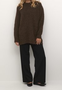 Brun, fluffig stickad tröja med lös passform och långa ärmar, ihop med högmidjade svarta vida byxor och platta svarta skor.