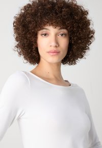 Kihara inimene, kes kannab valget pikakäist topi laia kaelusega. Materjal näib sile ja kerge. Neutralne taust.