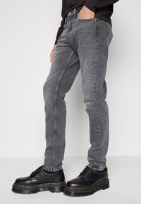 Jean en denim gris avec une coupe ajustée, doté de cinq poches et d'un dégradé subtil, associé à des chaussures en cuir noir avec des semelles en caoutchouc épaisses.