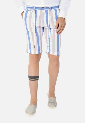 Uomo che indossa pantaloncini beige, bianchi e blu a righe verticali con una camicia bianca, scarpe slip-on grigie e un tatuaggio nero a doppio nastro sul polpaccio sinistro.