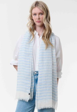 Blonde woman with wavy hair wears a white button-up shirt, light blue striped scarf, and denim jeans against a plain white background.