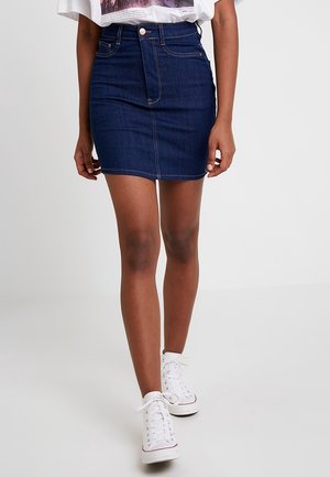 Kobieta ubrana w mini spódniczkę z ciemnoniebieskiego dżinsu, białą koszulkę z nadrukiem i białe tenisówki na sznurowadła, stojąca na tle jasnego, jednolitego tła.