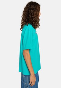 Person with long curly hair wearing a turquoise oversized t-shirt and blue cargo denim jeans standing in profile on a white background.