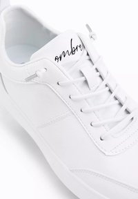 Sneaker bianco con lacci realizzato in materiale sintetico liscio. Presenta una punta arrotondata, cuciture decorative e un logo nero sulla linguetta.