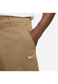 En mörkhyad mans hand i framlommen på beige Nike-byxor med vitt swoosh-logotyp, synlig midja och vit skjortekant ovanför.