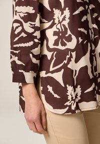 Langärmliges Hemd in Braun mit cremefarbener Blumenmuster, aus leichtem Stoff gefertigt. Verfügt über Knopfdetails und eine lockere Passform.