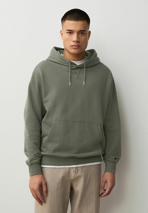 Young man wearing an olive green hoodie with drawstrings and beige pants, standing against a plain light gray background.