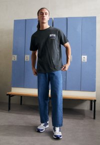 Dunkelgraues Baumwoll-T-Shirt mit "INTER" in Weiß und Gelb bedruckt, kombiniert mit blauen Jeans und grauen Sneakers mit blauen Akzenten.