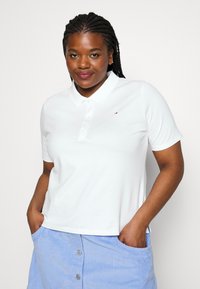 Camisa polo blanca con collar y tres botones, que presenta un pequeño logo bordado. Tejido suave con un corte holgado, combinada con una falda azul.