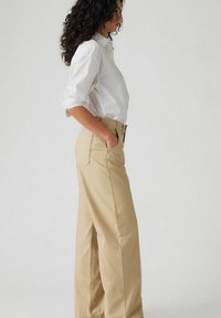 Beige wijde broek met zijzakken en een hoge taille, gecombineerd met een aansluitend wit shirt, weergegeven in profielzicht.
