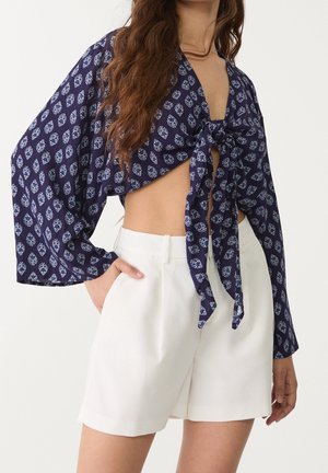 Kvinde iført en marineblå mønstret langærmet cropped top bundet foran og højtaljede hvide shorts med lommer.