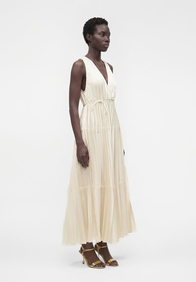 Femme portant une robe maxi plissée crème sans manches avec un col en V et une ceinture à la taille, associée à des sandales dorées à talons hauts, debout sur un fond uni.