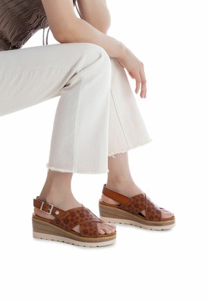 Sandalias de cuña de cuero marrón con patrón floral perforado, correa ajustable en el tobillo y una plataforma ligera de madera. Se llevan con pantalones blancos con flecos.