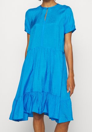 Woman wearing a short-sleeved, knee-length bright blue dress with a ruffled hem and small front neckline slit.